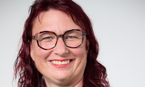 A portrait of a smiling woman wearing glasses, a dark blue shirt and a silver pendant necklace
