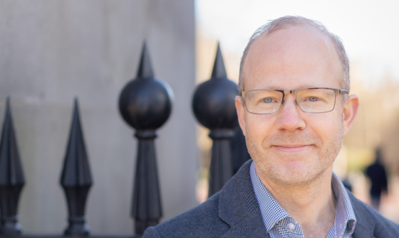 A man wearing glasses, blue shirt and jacket, standing in front of a wrought iron fence