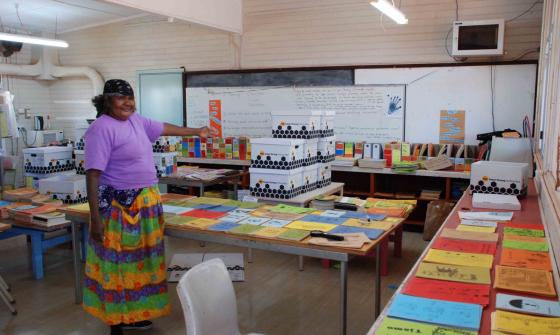 A woman stands in a classroom gesturing at piles of readers laid out on the tables in the classroom.