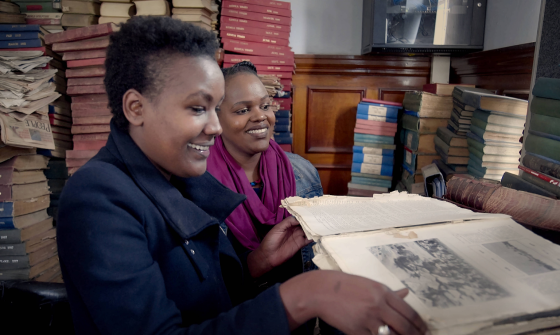 Two smiling women examining an open book with books piled high around them