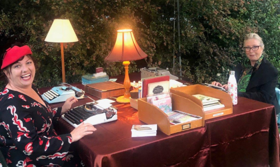 Two women are seated at a table covered in red cloth and laid with vintage lamps, stationery and typewriters