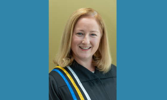 A blond-haired woman wearing courtroom robes with blue, yellow and white ribbons on the right shoulder