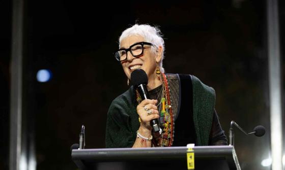 A woman with white hair and dark-rimmed glasses is speaking into a microphone