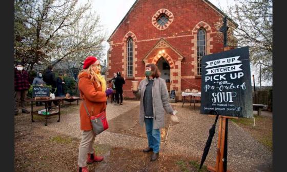 People gathering outside a red brick church, with a sign advertising a 'Pop-Up Kitchen' and soup pickup.