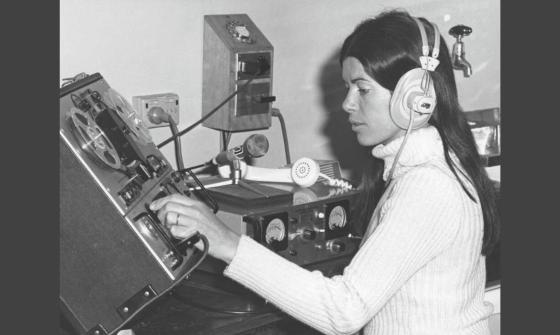 A black and white photo of a woman wearing headphones, operating a vintage audio recording or mixing console in a studio setting.