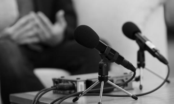 Black and white image of recording equipment and microphone on desk, with a pair of hands in the background