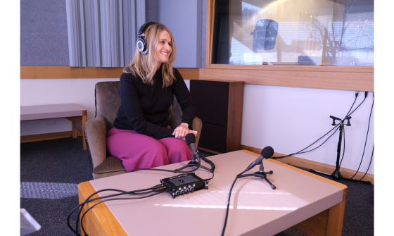 A smiling blonde woman wearing headphones is seated in a chair in a modern studio setting, facing a desk with audio equipment and a microphone setup.