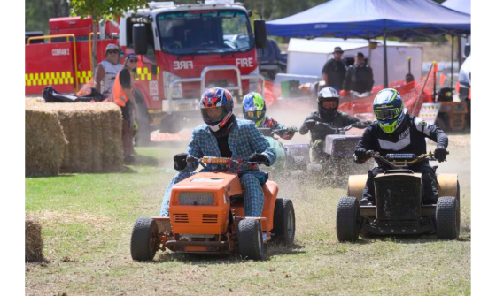 Four individuals race ride-on lawn mowers on a grassy field. A fire truck is visible in the background.