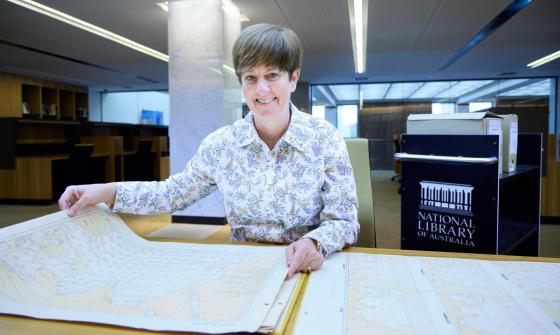 A smiling woman with short brown hair sits at a desk, holding open a large, detailed historical map. She wears a floral-patterned blouse and looks directly at the camera..