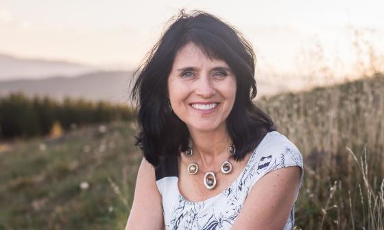 A woman with dark, wavy hair sitting outdoors, wearing a white patterned top.
