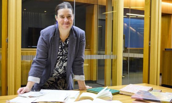 A woman with dark hair pulled back smiles at the camera while leaning on a wooden table. She wears a grey blazer over a patterned blouse. Several books and open notebooks are spread across the table in front of her.