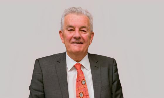 A smiling man with grey hair wearing a grey suit, white shirt and a red patterned tie with circular emblems. He is photographed against a plain light gray background.