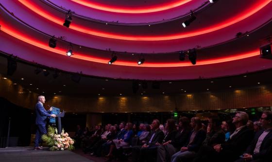 A man in a suit speaks at a podium on a stage in a modern auditorium with a striking red-lit circular ceiling. An audience sits in rows, attentively listening.
