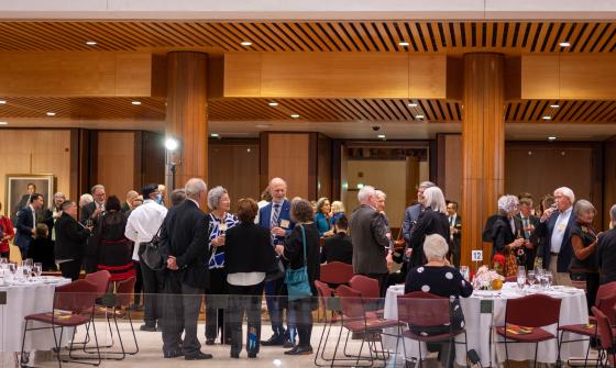 A group of adults in formal attire mingle and converse in a spacious, wood-paneled event hall with round tables set for dining.