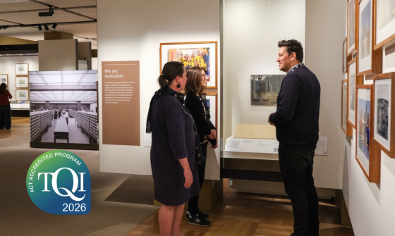 Three people are viewing exhibits in a museum gallery. A man stands facing two women, appearing to explain something about displayed artwork and documents. A large informational panel titled 'We are Australian' is visible, along with framed pictures on the walls and a display case. A 'TQI ACT Accredited Program 2026' logo is in the lower-left corner.