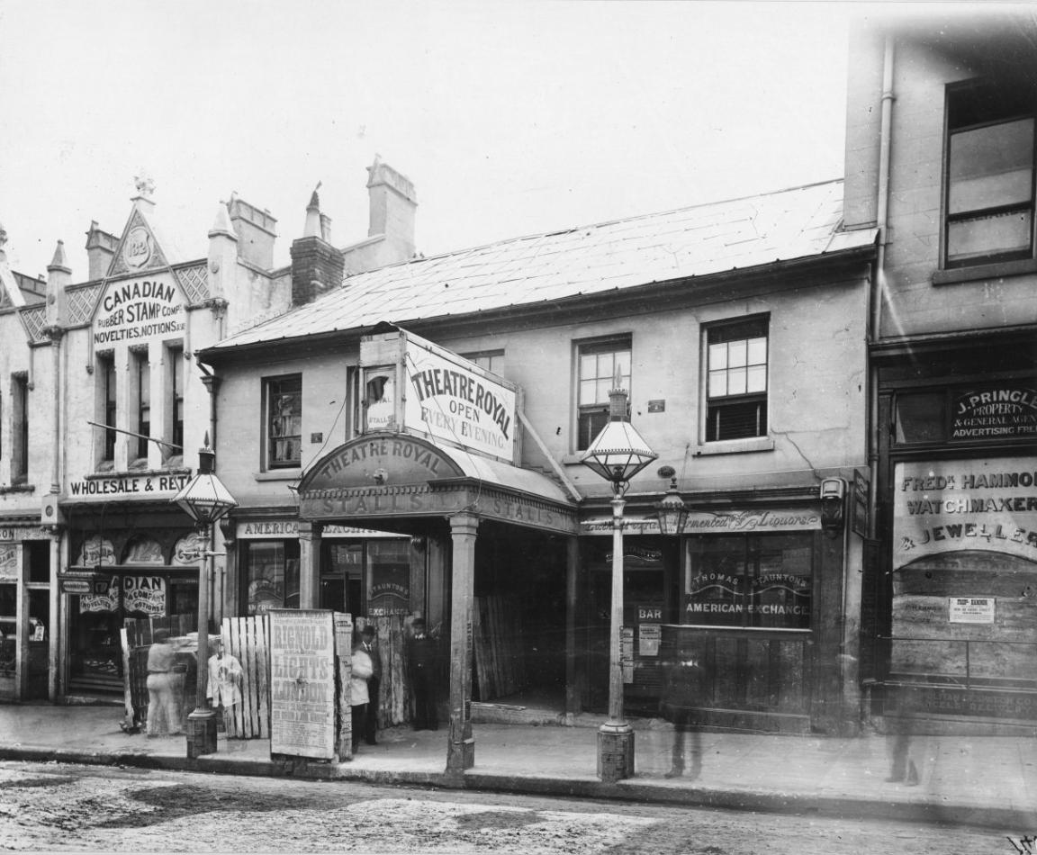 Black and white photograph of a row of shopfronts. One facade reads 'Theatre Royal: Open Every Evening'.