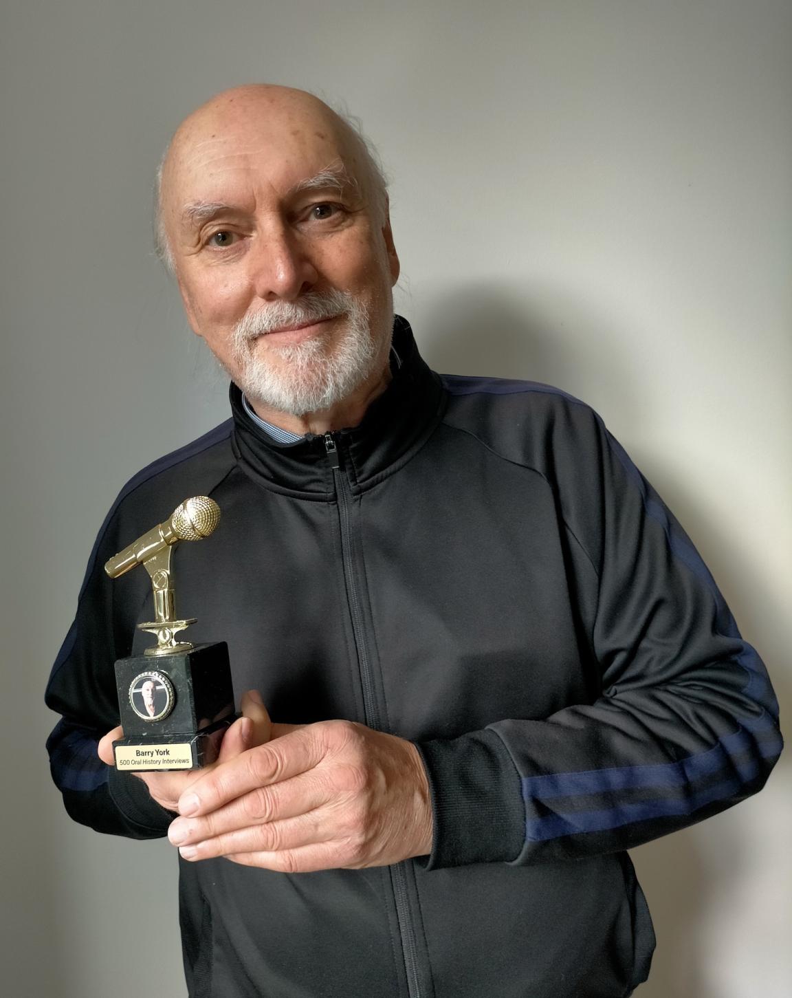 Middle-aged man with a white beard smiling and holding up a trophy with a golden microphone that reads 'Barry York 500 Oral History Interviews'