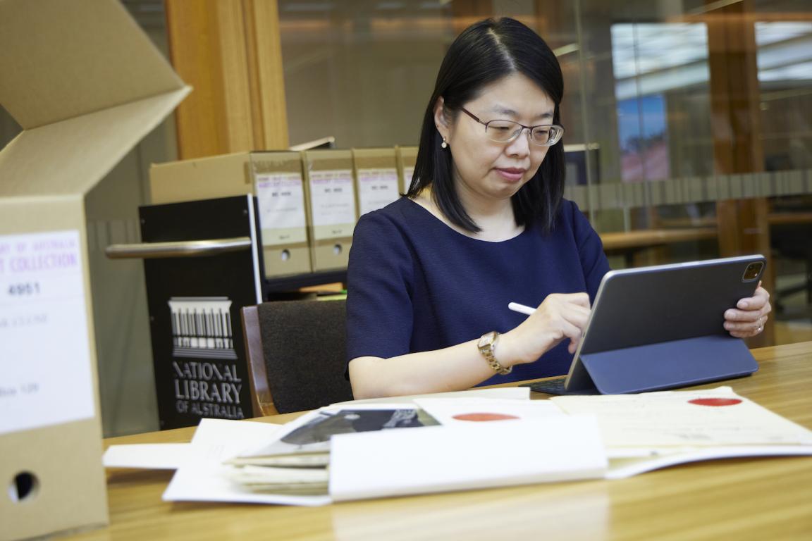 Woman with black hair and wearing a navy top using a laptop with a stylus at a desk covered in papers and a box