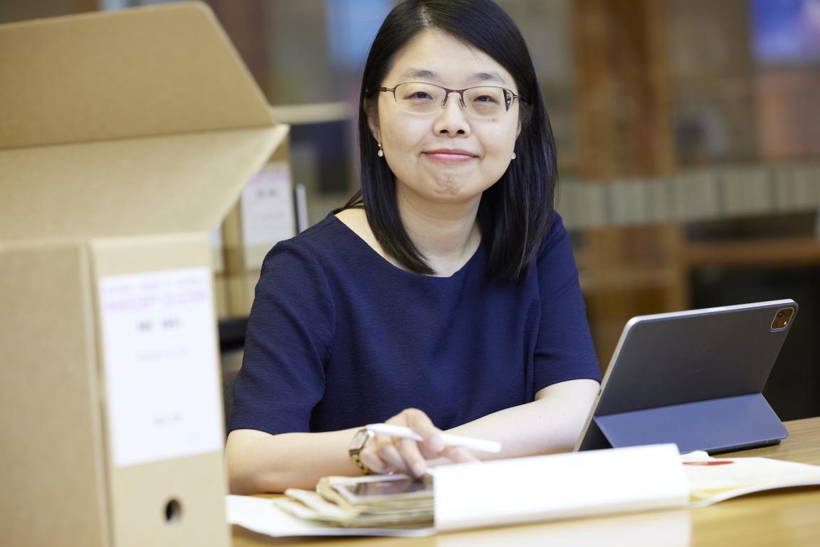 Woman with short dark hair wearing a navy short sleeve top smiling at the camera and sitting at a desk with a laptop, papers and a box