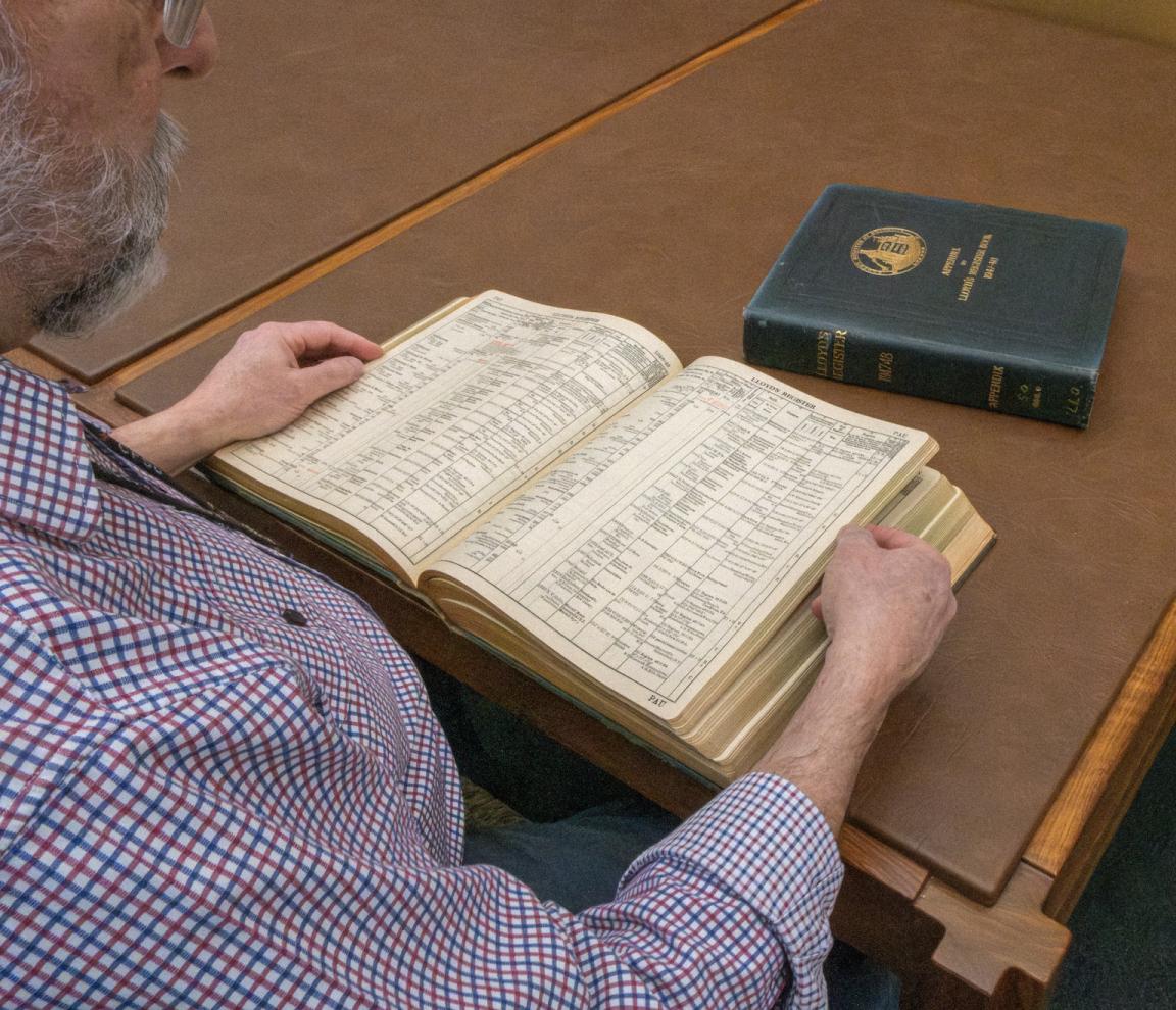Man reading through a thick, leather-bound book of shipping registers