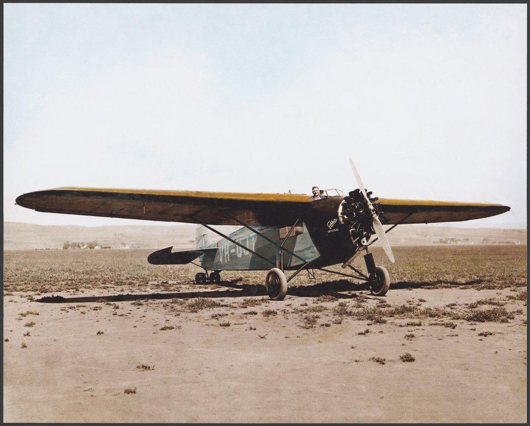 An old plane on the ground in a desert. A man is poking his head out from the cockpit and can be seen behind the wings of the plane