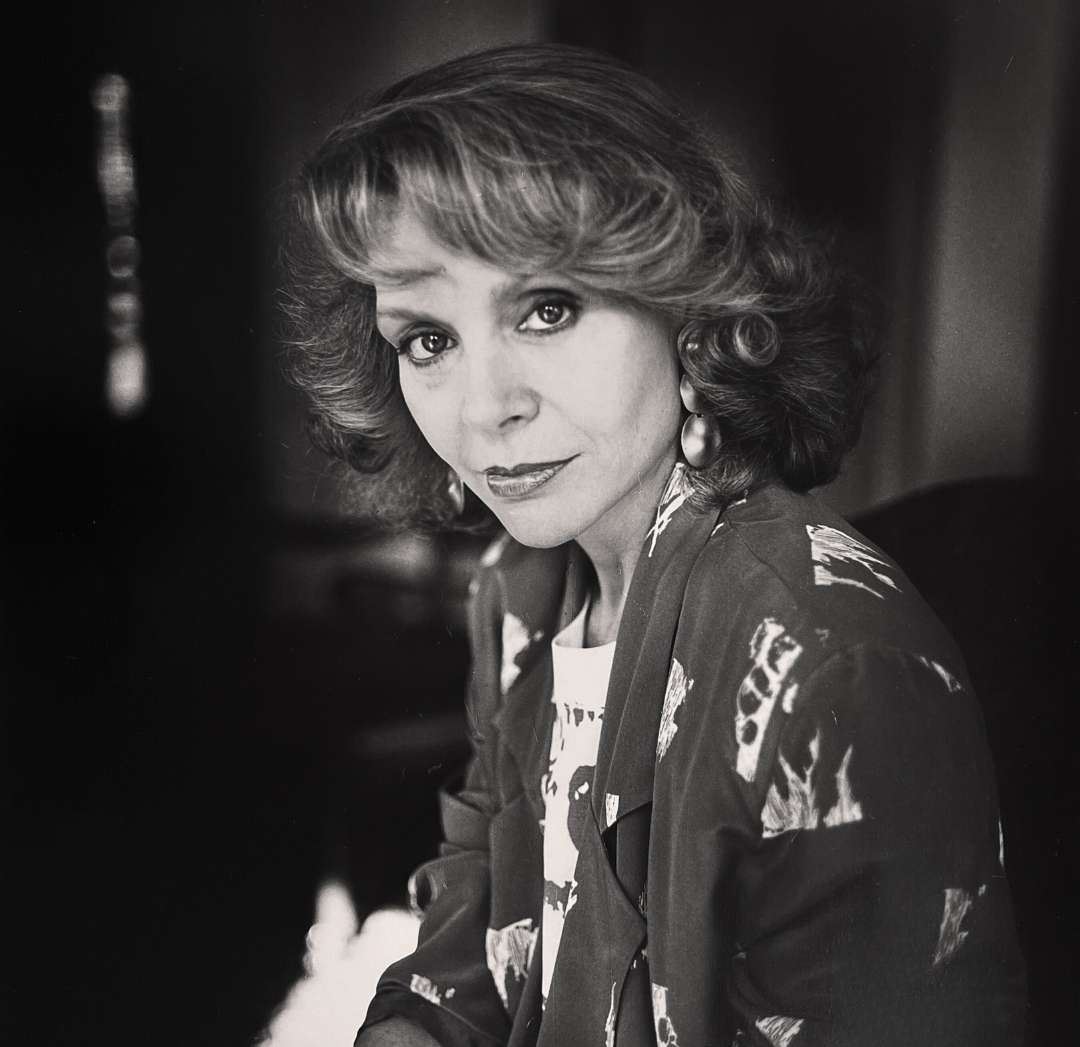 Black and white photograph of a woman with light, short curly hair looking directly into the camera with a small smile. She's wearing a dark blazer with light, abstract shapes on it and large round earrings
