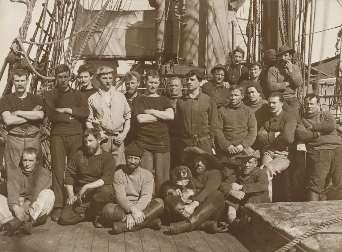Sepia toned photo of a chips crew made up of about 20 men and a dog, on the deck of their ship 'Terra Nova'