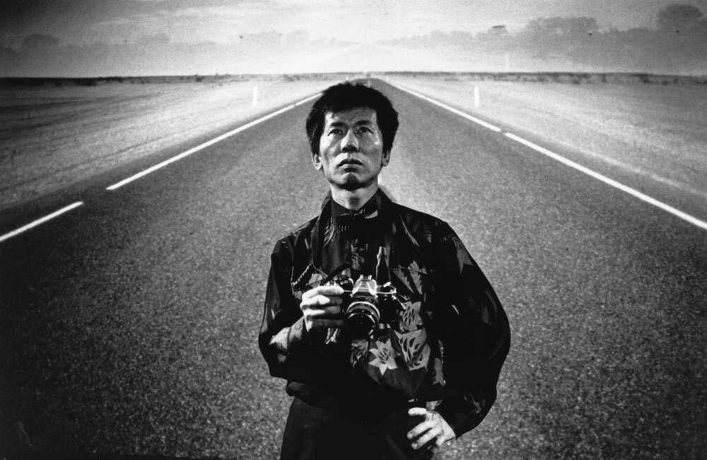 A black-and-white portrait of William Yang standing alone on an empty road, holding a camera. The road stretches behind Yang as he gazes straight into the camera in a solemn pose.