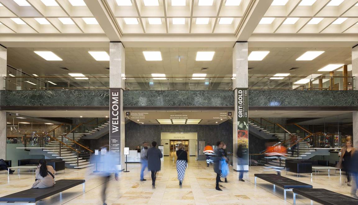 Large two-story room with cream-coloured marble floors, columns and stairs; black leather couches; lots of light from the ceiling and a balcony from the second floor looking over the room. On the columns are signs with directions and the word 'Welcome'. People are moving around the room.