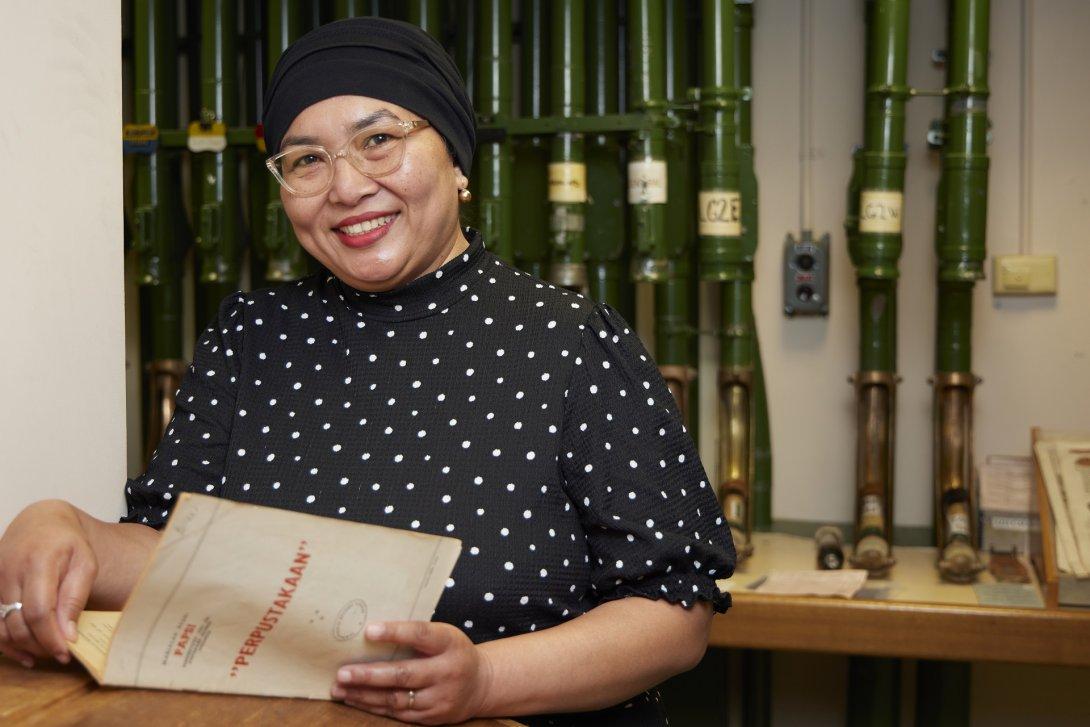 Woman wearing a dot print shirt, glasses and a black head scarf holding an open booklet and smiling