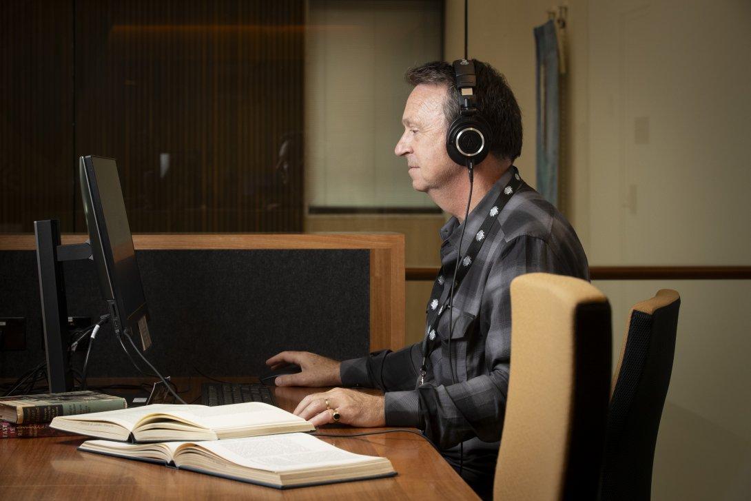 Man in a grey and black checkered shirt and headphones sitting at a desk, using a computer. To his left are two open books