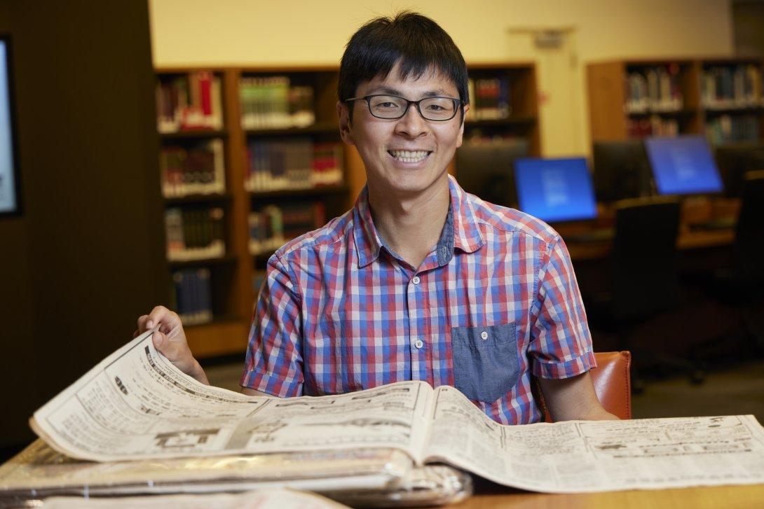 Man with short black hair and black glasses in a short-sleeved checkered shirt smiling and sitting at a desk with a Chinese language newspaper on it