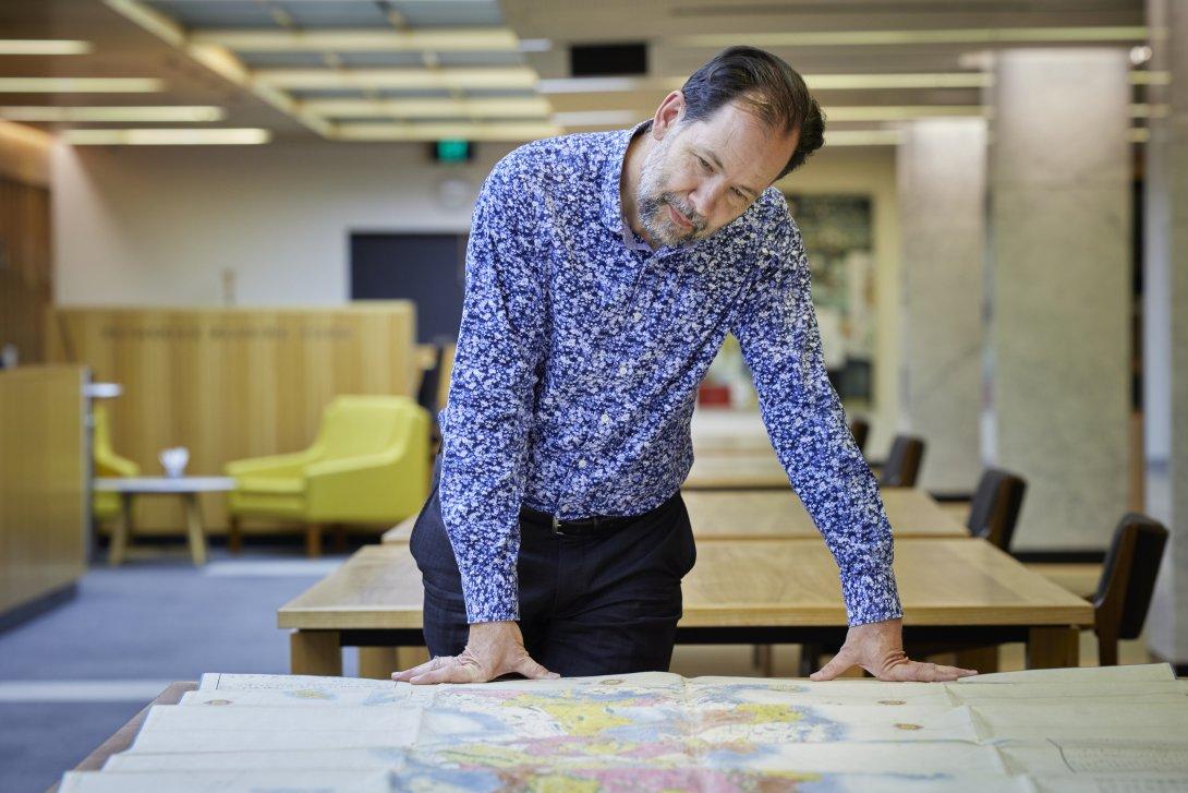 Man with brown hair and a grey and brown beard sitting at a table with a large map spread over it