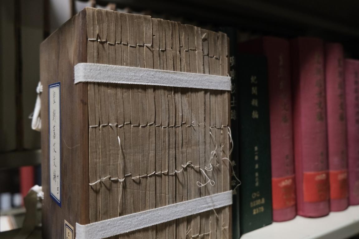 A large, old text with Chinese characters is bound with traditional thread stitching, sitting on a shelf, supported by a white fabric strap wrapped around its cover. The edges of the pages are uneven, showing wear with the stitching slightly frayed. Other colourful books with Chinese characters are lined up alongside it on the shelf.