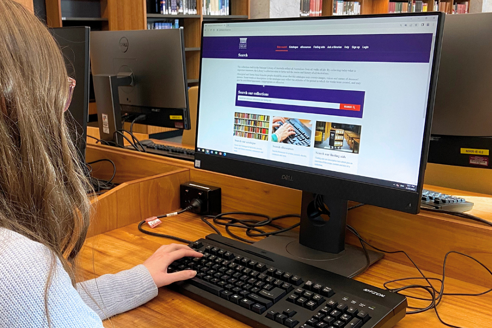 Young woman in blue knit jumper working at computer with the National Library's redeveloped catalogue on the screen