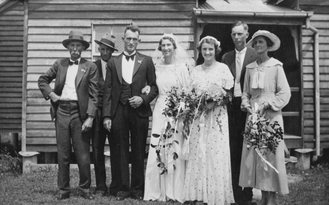 Smiling bride and groom with five family members standing in front of a small building