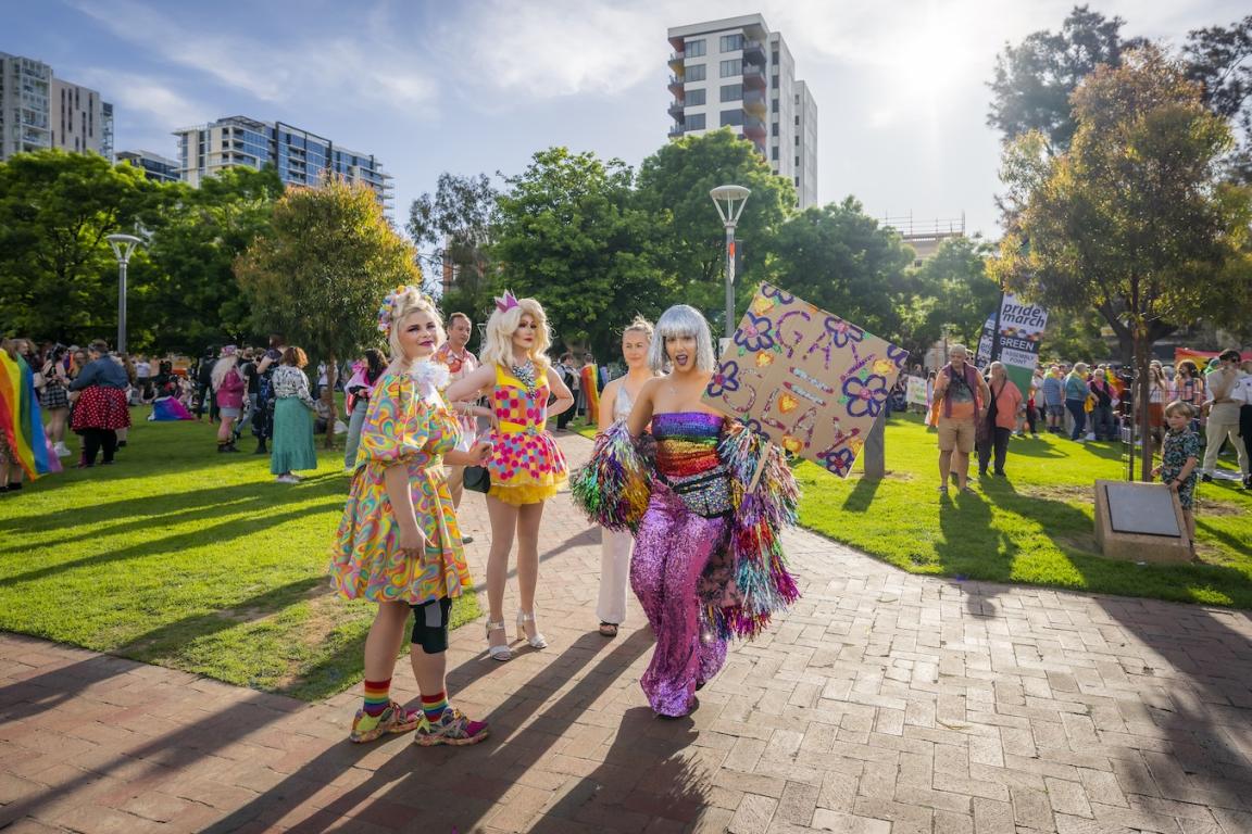 A group of four people who are very colourfully dressed, holding signs in a park. There are more groups of colourful people in the background.