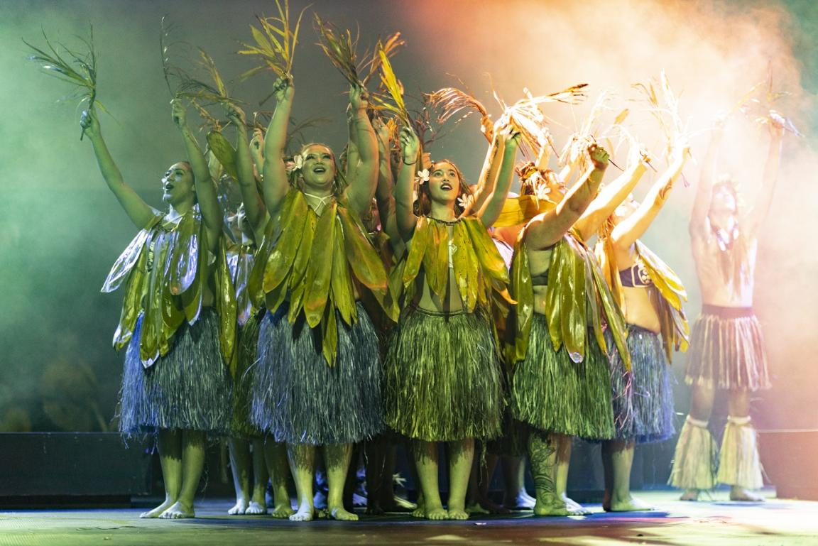 Group of dancers wearing large leaves around their necks and grass skirts, standing in a circle with their arms raised