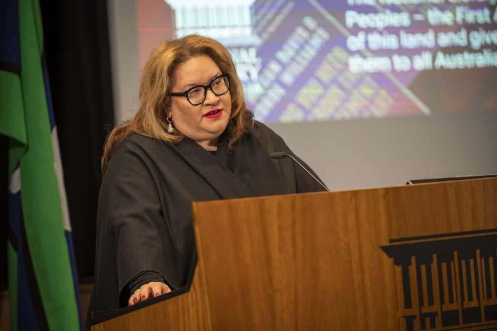 A person with long brown hair and glasses stands behind a wooden lectern, speaking into a microphone. The background features a screen displaying partially visible text and graphics. To the left, green and blue from the flag of the Torres Strait Islands is visible.