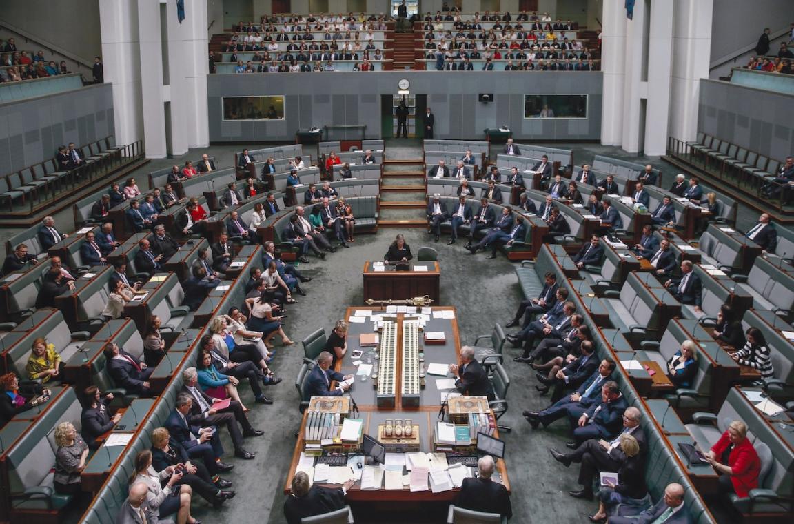 View of the Australian House of Representatives in Parliament House from slightly above. Members of the House of Representatives sit throughout the room, and members of the public sit in allocated areas