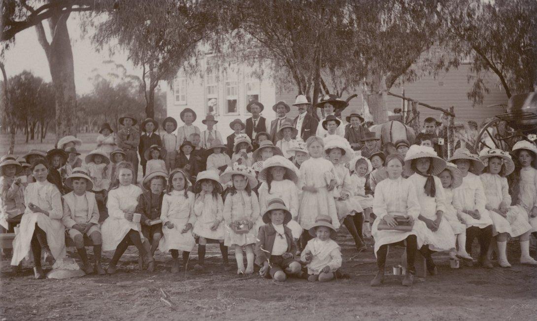 School portrait with dozens of young children sitting and standing under trees near the school building