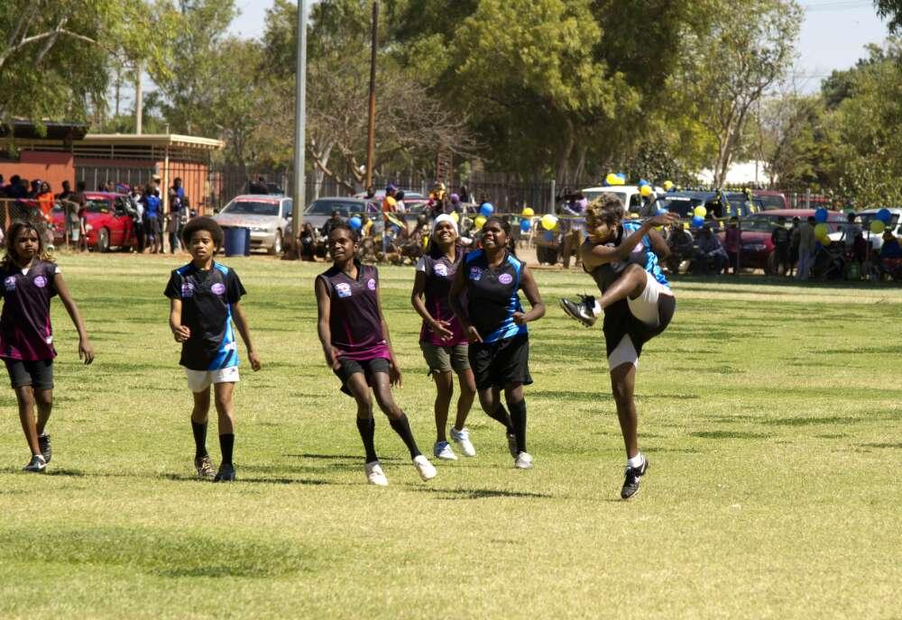 Six women smiling as they play football. One has just kicked the ball.