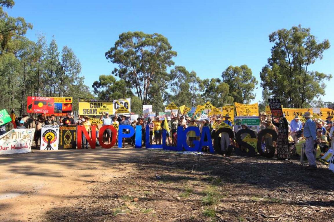 A group of protestors gathers at the entrance of a rural site, holding signs and banners against coal seam gas (CSG) mining. The large text “NO PILLIGA CSG” is displayed in colorful letters, with people standing behind, surrounded by trees and a clear sky.