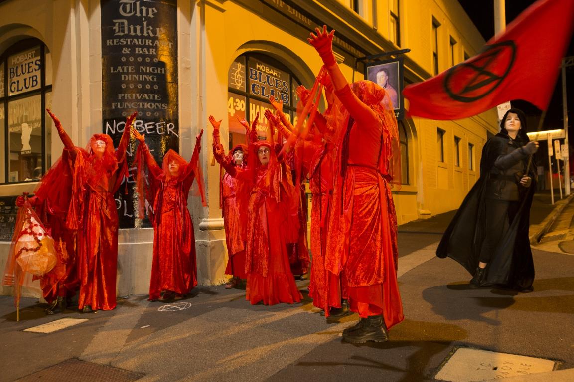 "A group of Extinction Rebellion Red Brigade protesters dressed in flowing red costumes and veils stand with arms raised in a dramatic gesture on a dimly lit street corner in Hobart, Tasmania. One protester, holding a large red flag with an extinction symbol, stands slightly apart. The backdrop includes a bar and club with signs reading 'NO BEER' and 'Hobart Blues Club.'