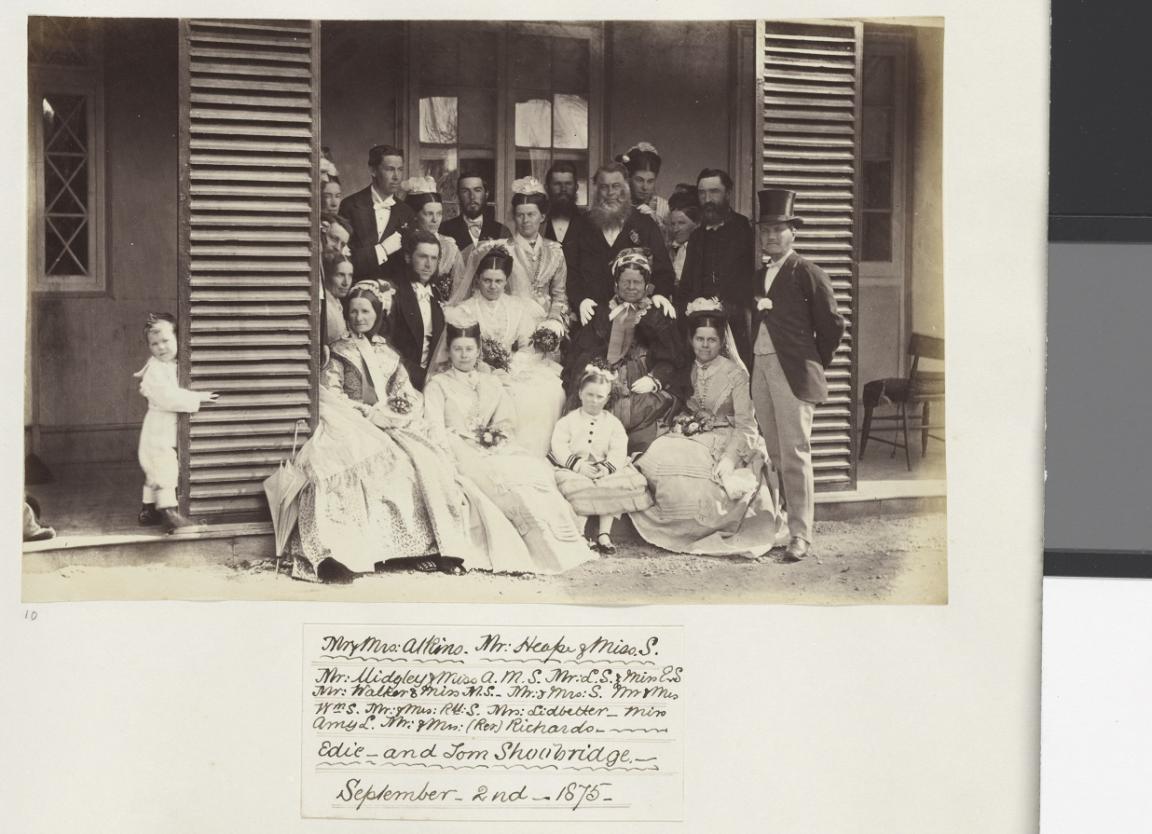 A black-and-white photograph showing a large wedding party posed on the steps of a wooden house. The group includes men, women, and children dressed in formal Victorian attire. The women are in elaborate dresses, some holding small bouquets. The men wear suits, some with top hats, and a small boy peeks around a door frame to the left.