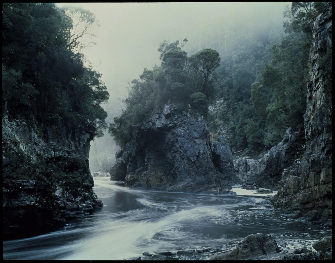 A tranquil river bends through a rugged gorge surrounded by dense forest. The rocky cliffs, covered with lush vegetation, rise steeply on both sides, while morning mist creates a soft, hazy atmosphere. The water, smooth and glassy, swirls in the foreground, capturing the calm yet powerful essence of the scene. The landscape evokes a sense of natural beauty and wilderness.