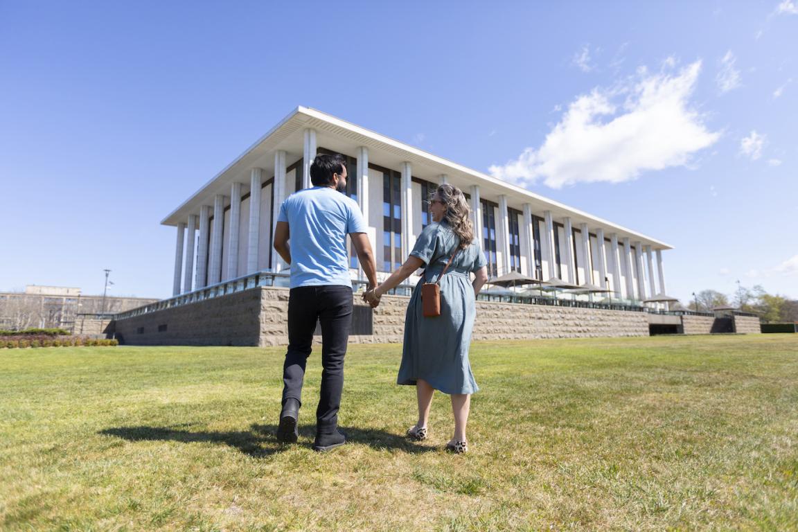 Man and woman in casual wear holding hands and laughing as they walk towards the National Library building