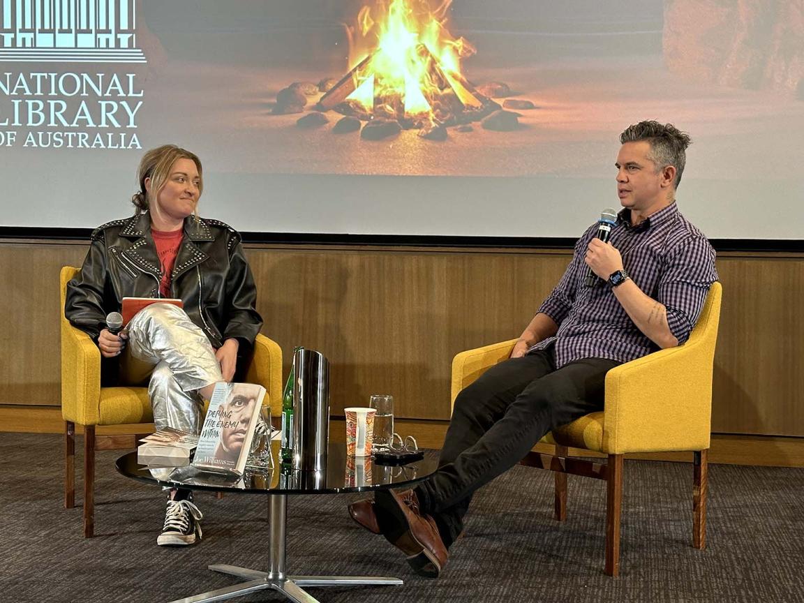 A man and a woman sit in chairs on stage with a picture of a campfire behind them. The woman holds a microphone and the man is speaking into a microphone.