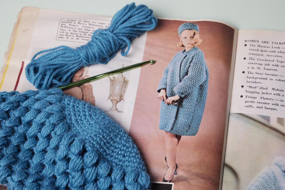 An old magazine showing a vintage crochet pattern. Laying on top of the magazine is a blue crochet item in progress.