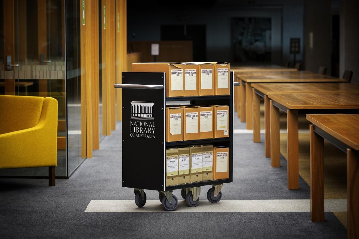 A National Library of Australia archival cart with labeled special collections boxes, placed in a reading room beside wooden tables and yellow seating.
